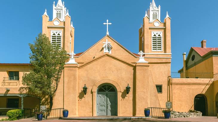 A cathedral with sandstone walls, two towers, and a cross atop it next to a tree A cathedral with sandstone walls, two towers, and a cross atop it next to a tree