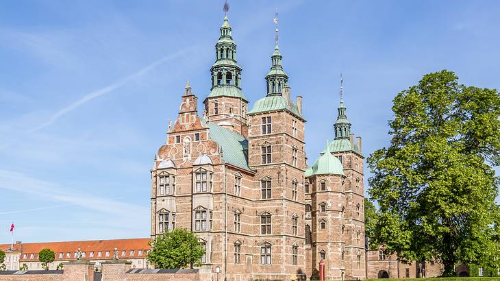 A renaissance-style brick castle with spires next to green trees and a blue sky