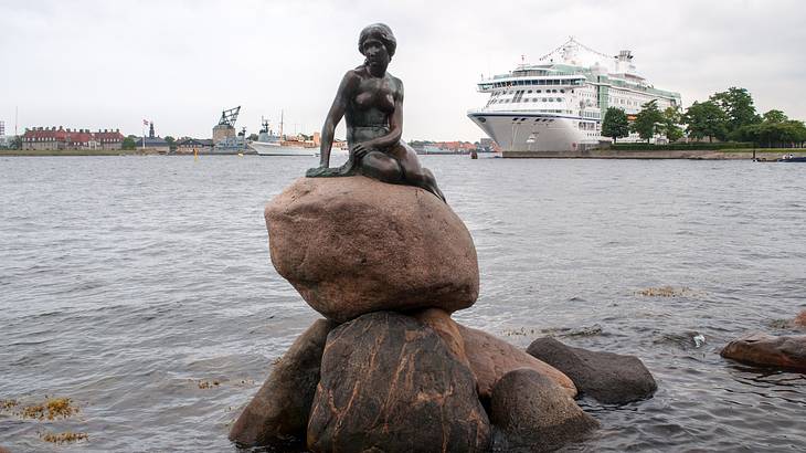A statue of a girl sitting on a large rock along the shore with a ship at the back