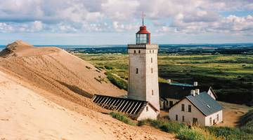 A lighthouse tower with a sand dune and a few structures surrounding it