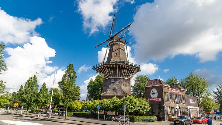 A tall wooden windmill next to green trees and a road under a blue sky with clouds