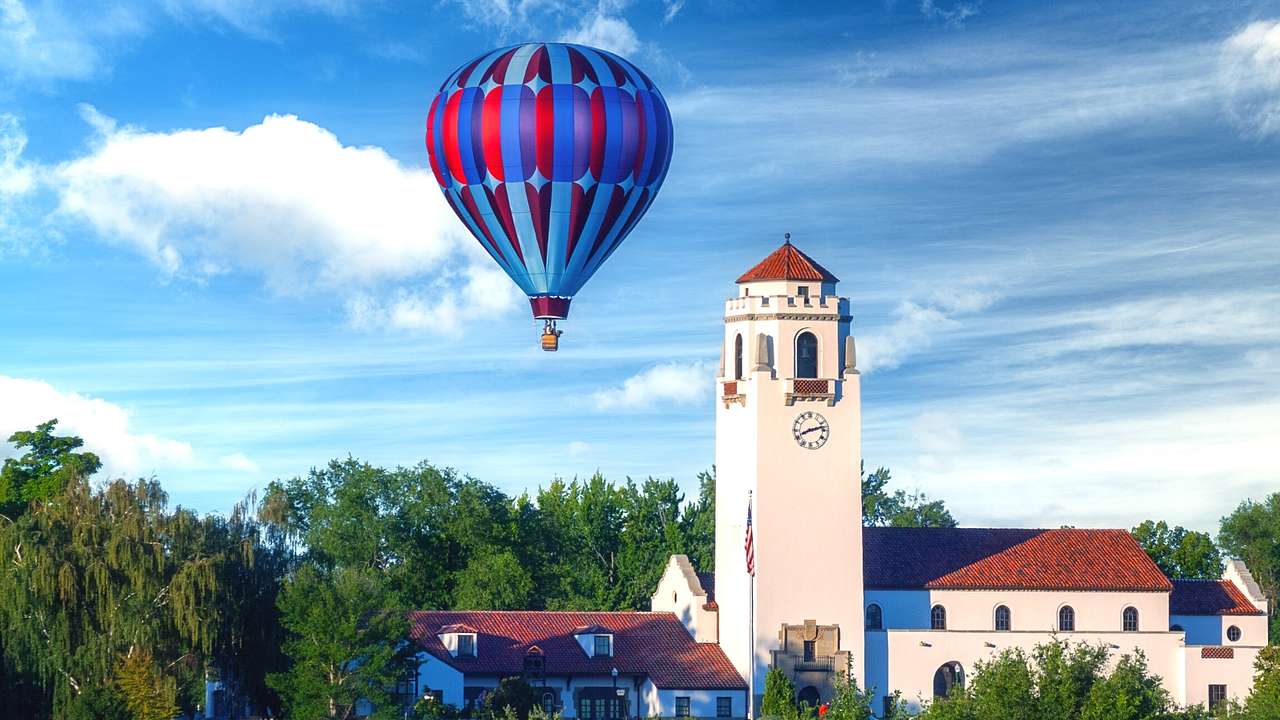 A hot air balloon in the sky next to trees and a white building with a clock tower
