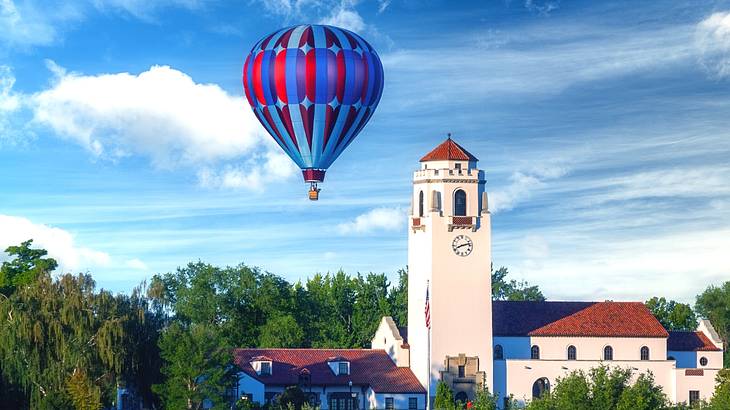 A hot air balloon in the sky next to trees and a white building with a clock tower