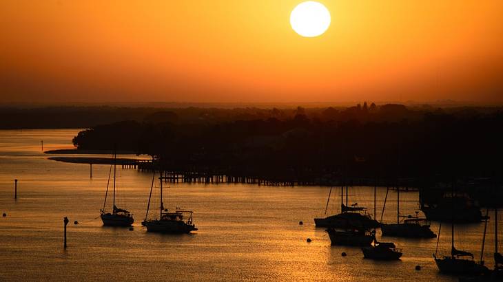 Several boats on water against a silhouette of a town under a yellow sky with the sun