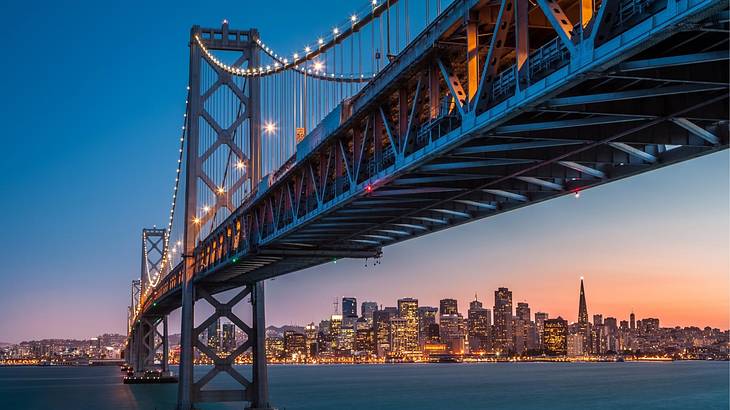 A suspension bridge over a bay with a skyline in the distance at night