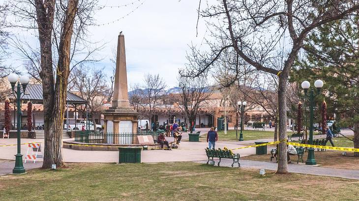 Local people hanging out on a square with a tall monument, trees, grass, and benches Local people hanging out on a square with a tall monument, trees, grass, and benches