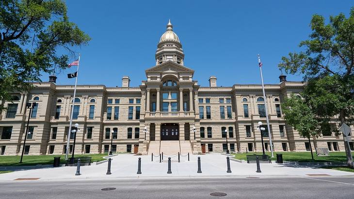 A large state capitol building with a domed roof and steps, flags, and trees in front A large state capitol building with a domed roof and steps, flags, and trees in front