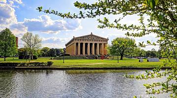 A Parthenon in a park with trees, green grass, and a lake in front of it