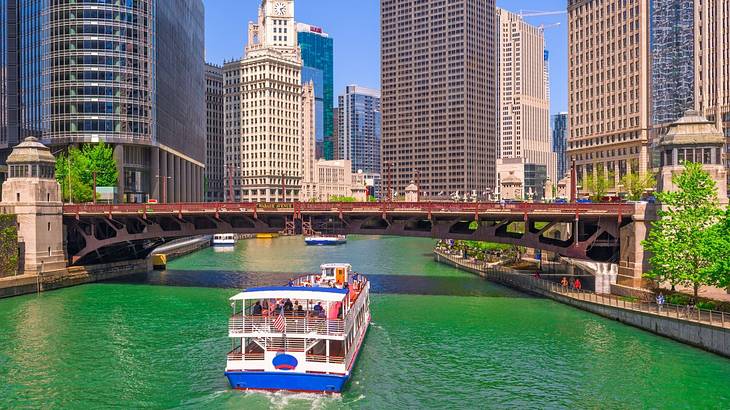 A small cruise boat on a river going toward a bridge with buildings surrounding