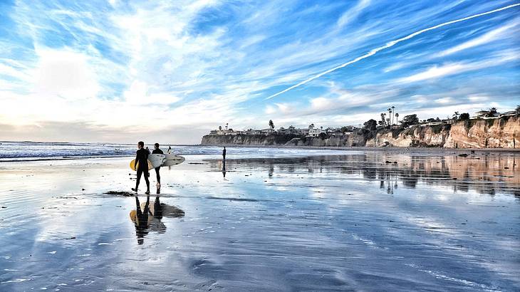 Two people with surfboards walking on a beach with wet sand