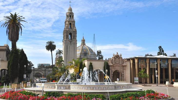 A garden with a fountain and palm trees and a building and stone tower behind