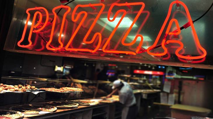 A red neon sign that says Pizza in front of a kitchen with pizza on the counter
