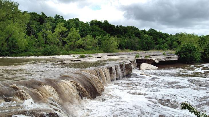 A small waterfall flowing into a pool with green trees behind it under a cloudy sky A small waterfall flowing into a pool with green trees behind it under a cloudy sky