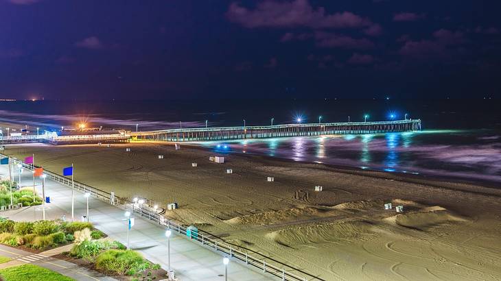A sandy beach next to a boardwalk and the ocean and a pier at night