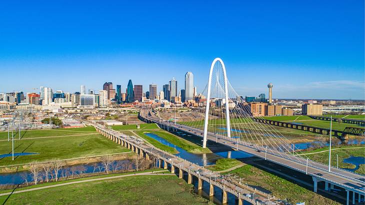 A city skyline next to grass and a white arch under a blue sky