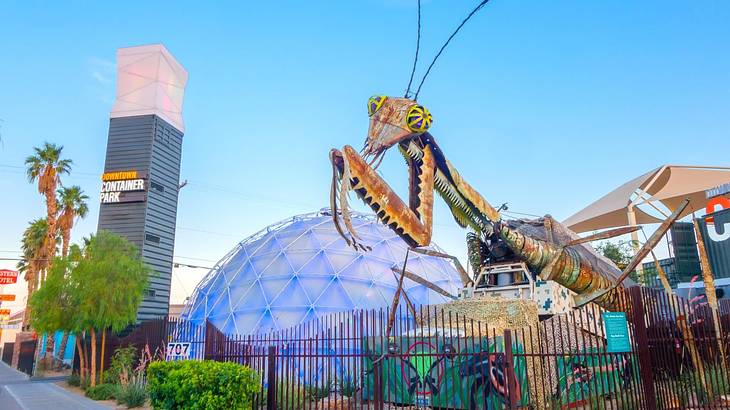 A praying mantis statue with a dome structure and other buildings behind it