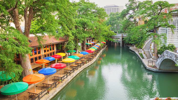 A river with a bridge on one side and trees and colorful patio umbrellas on the other