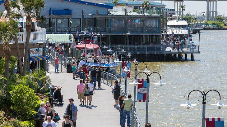 Crowds walking on a sidewalk next to a body of water with restaurants on the water