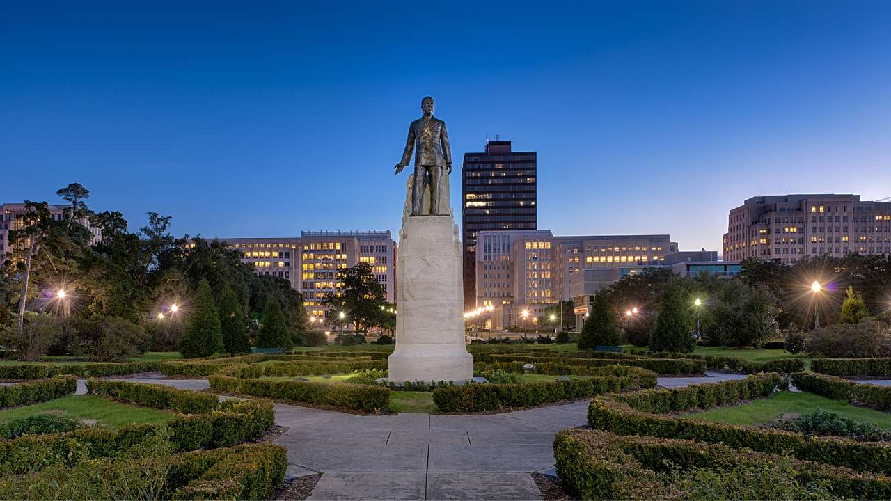A statue of a man surrounded by a path and garden next to city buildings at night