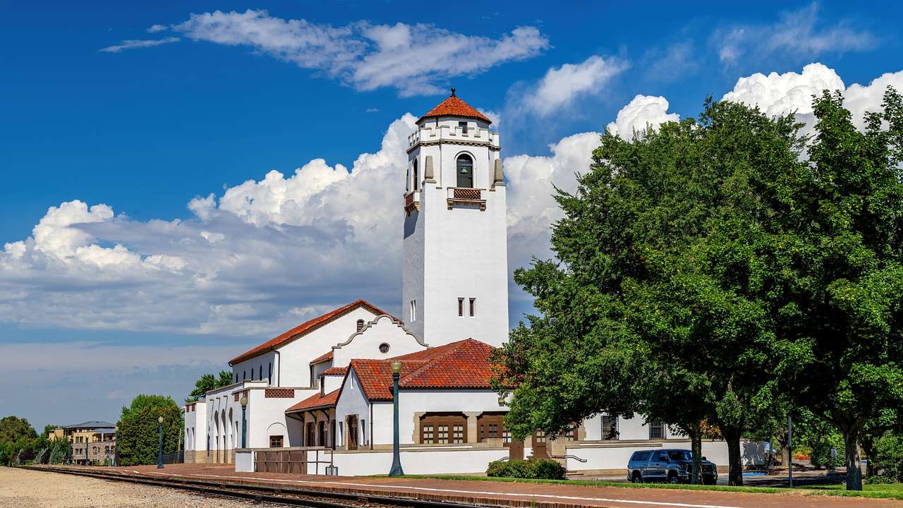 A white train building with a tower and orange roofs next to a train track and trees