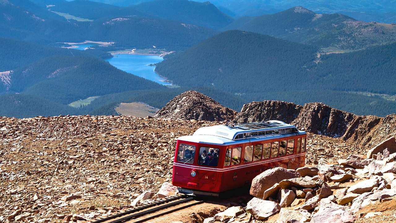 A small red train on a track on a mountain with a valley and lake below