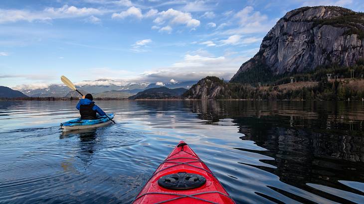 A red kayak in the foreground and a man in a kayak on the left side on calm water