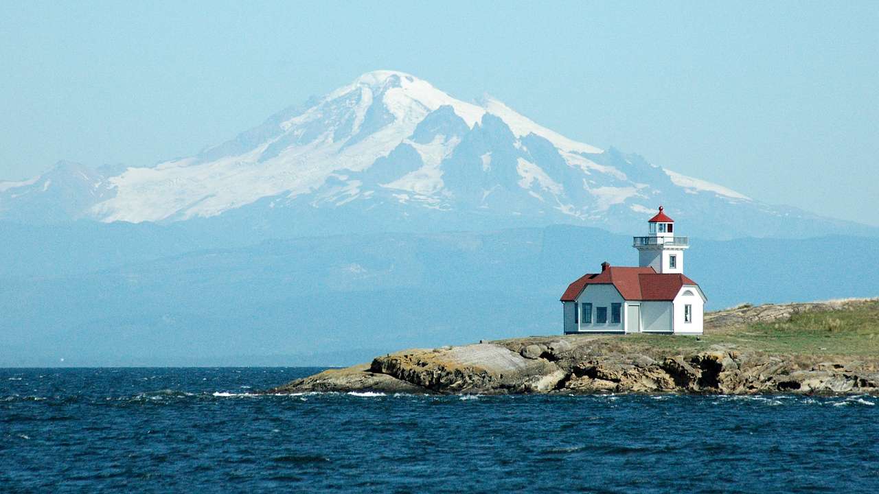 A white and red lighthouse on rocky green land, in front of a snow-capped peak