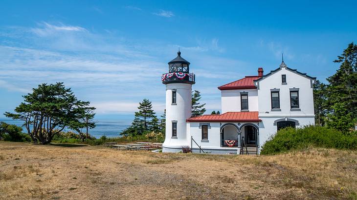 A white lighthouse behind brown grass with greenery around on a nice day