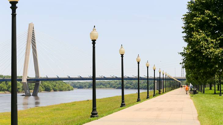 A walkway and lampposts beside a river with a bridge in the background