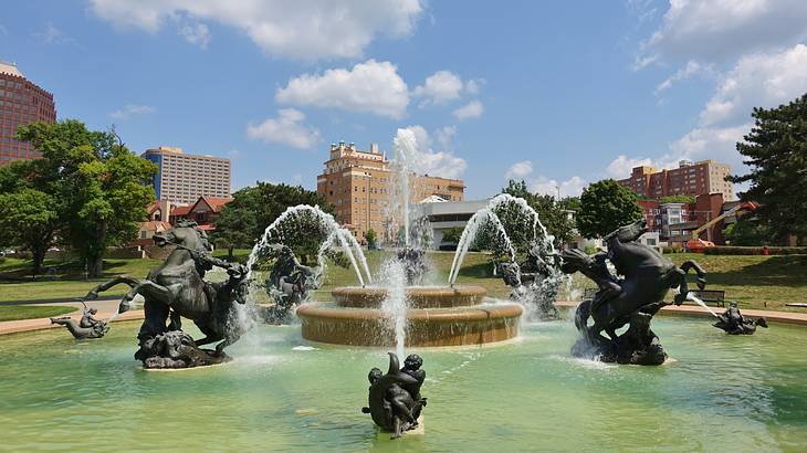 A fountain with sculptures of men riding horses next to trees and buildings