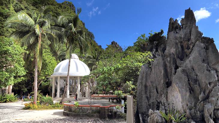 A white shrine surrounded by green trees and a tall rock on the right on a nice day