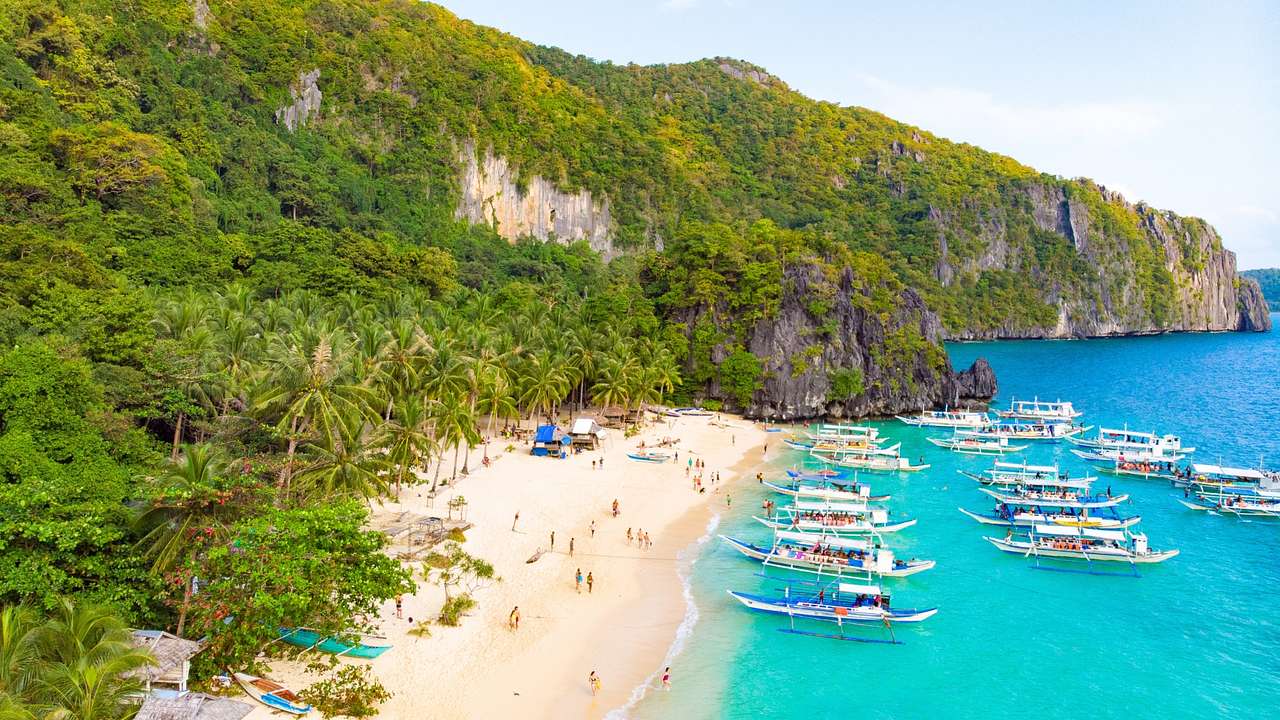 A white sand beach next to greenery-covered cliffs and turquoise water with boats