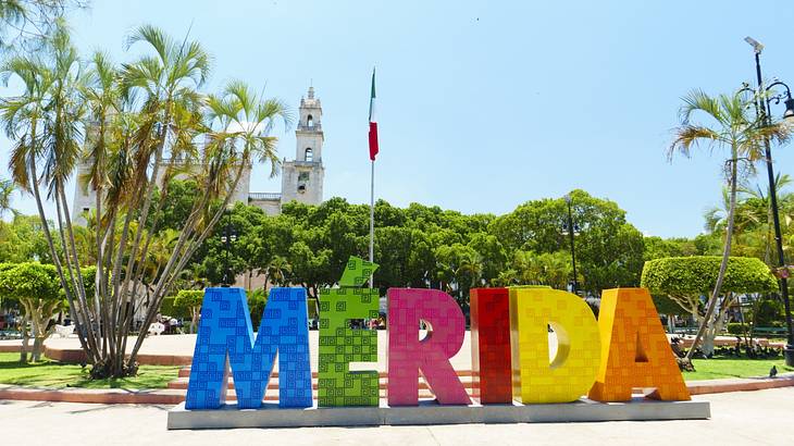 A colourful sign that says "Mérida" next to green trees and a white tower