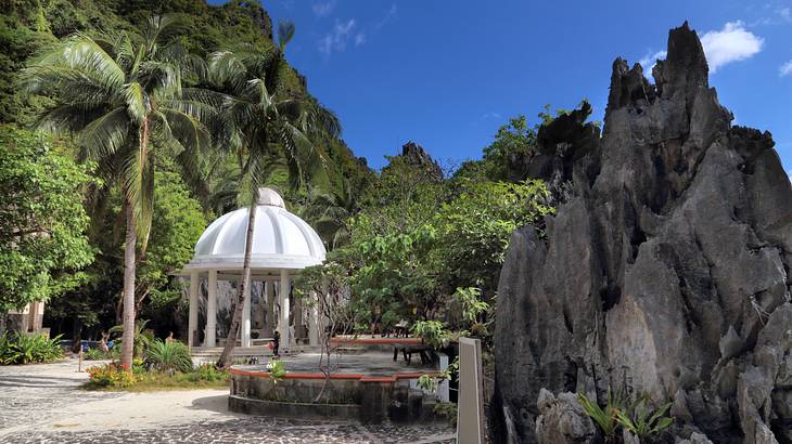 A white shrine surrounded by green trees and a tall rock on an island, Palawan