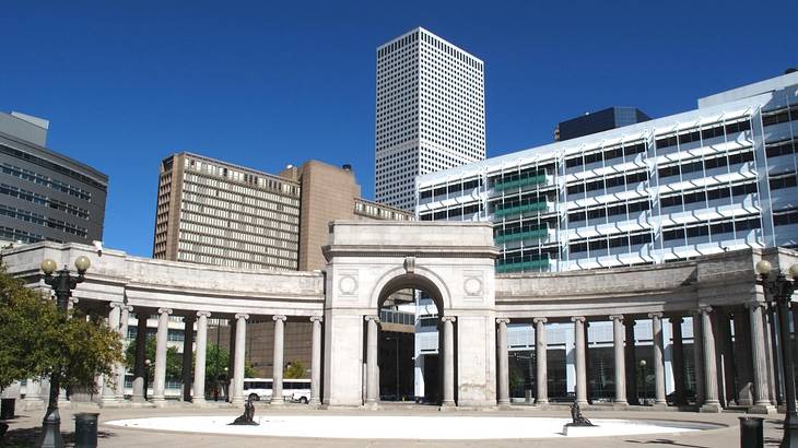 A Greek-style arch with tall buildings behind it under a blue sky