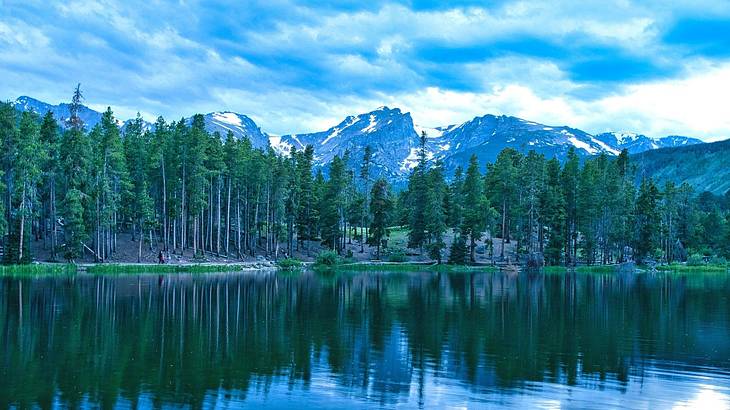 A green lake with mountains and green trees surrounding it under a blue cloudy sky