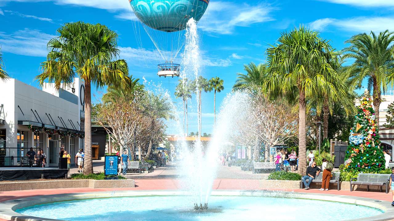 A fountain with a tree-lined street and shops at the back and a hot air balloon above