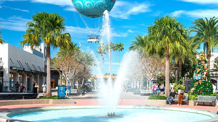 A fountain with a tree-lined street and shops at the back and a hot air balloon above