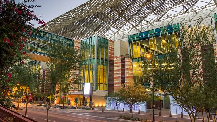 An illuminated modern glass-structure building with a road and trees in front