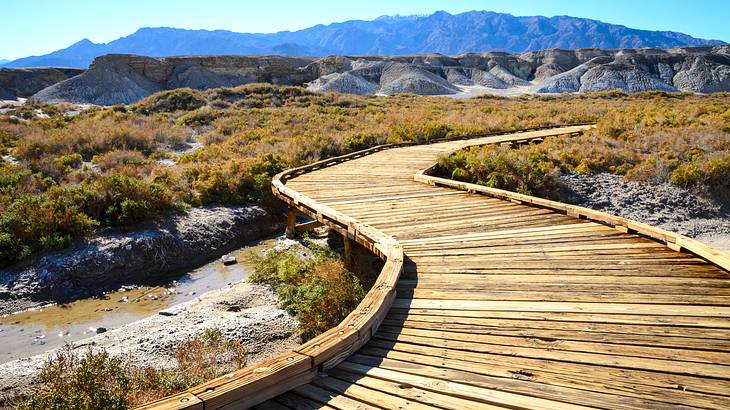 A wooden boardwalk in dry vegetation and a little creek and dry hills in the distance