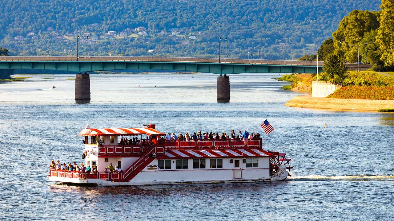 A red and white paddle-wheel riverboat on the water next to a bridge and greenery