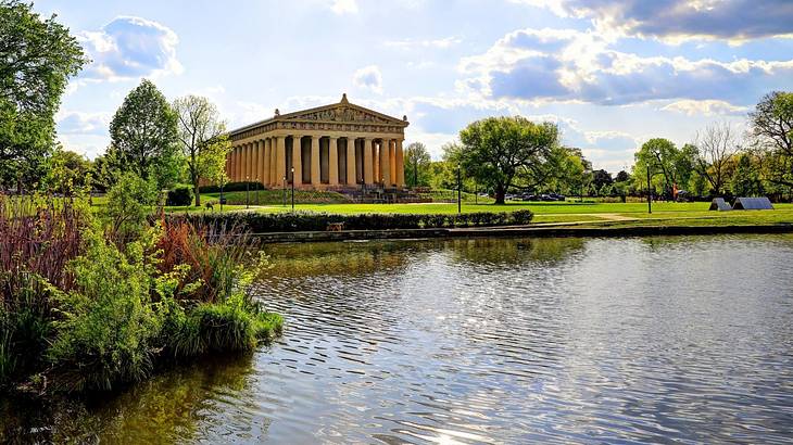 A Greek-style structure with a lake and greenery in front of it on a sunny day