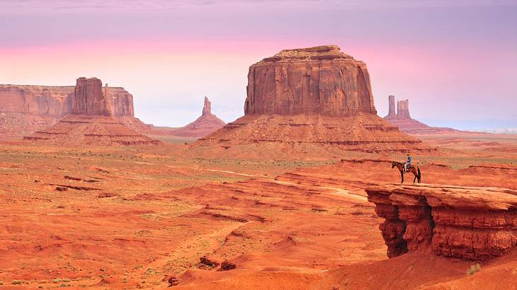 Red sandstone formations and a person on a horse on a cliff under a pink sunset sky