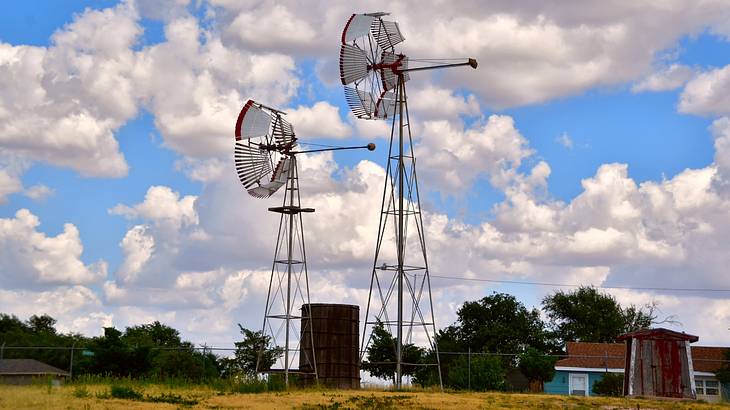 Two American windmills on the grass next to trees and a blue sky with clouds