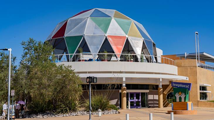 A dome-shaped building with colorful panels next to a path and a tree A dome-shaped building with colorful panels next to a path and a tree