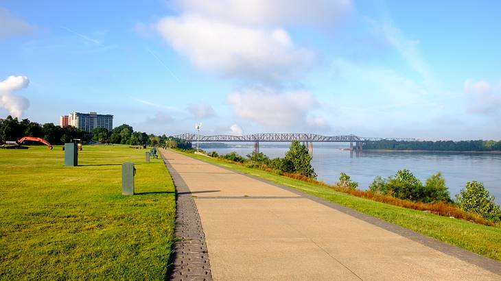 A paved walkway by a body of water and a lawn with a bridge in the distance