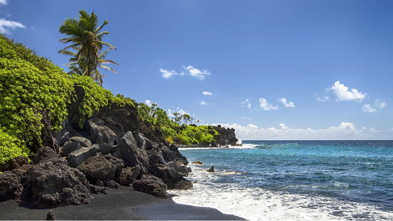 A black sand beach with lush trees and foliage in the distance on a partly cloudy day