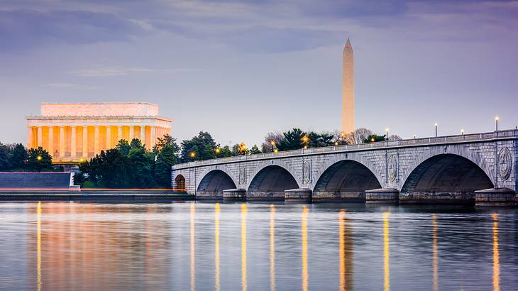 A river with a bridge, a building with columns, and an obelisk behind it at night