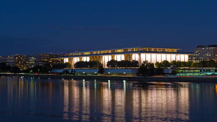 A rectangular building at night with the lights on and water in front of it