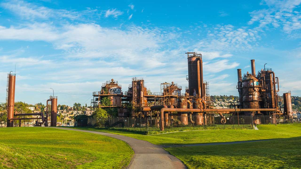 A park with a path and old iron gas plants under a blue sky with clouds